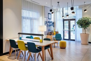 A modern hotel lobby with a dining area featuring a white table and colorful chairs, a large potted plant, and a view of the street through the glass doors. - Holiday Inn Nice by IHG (Nice)