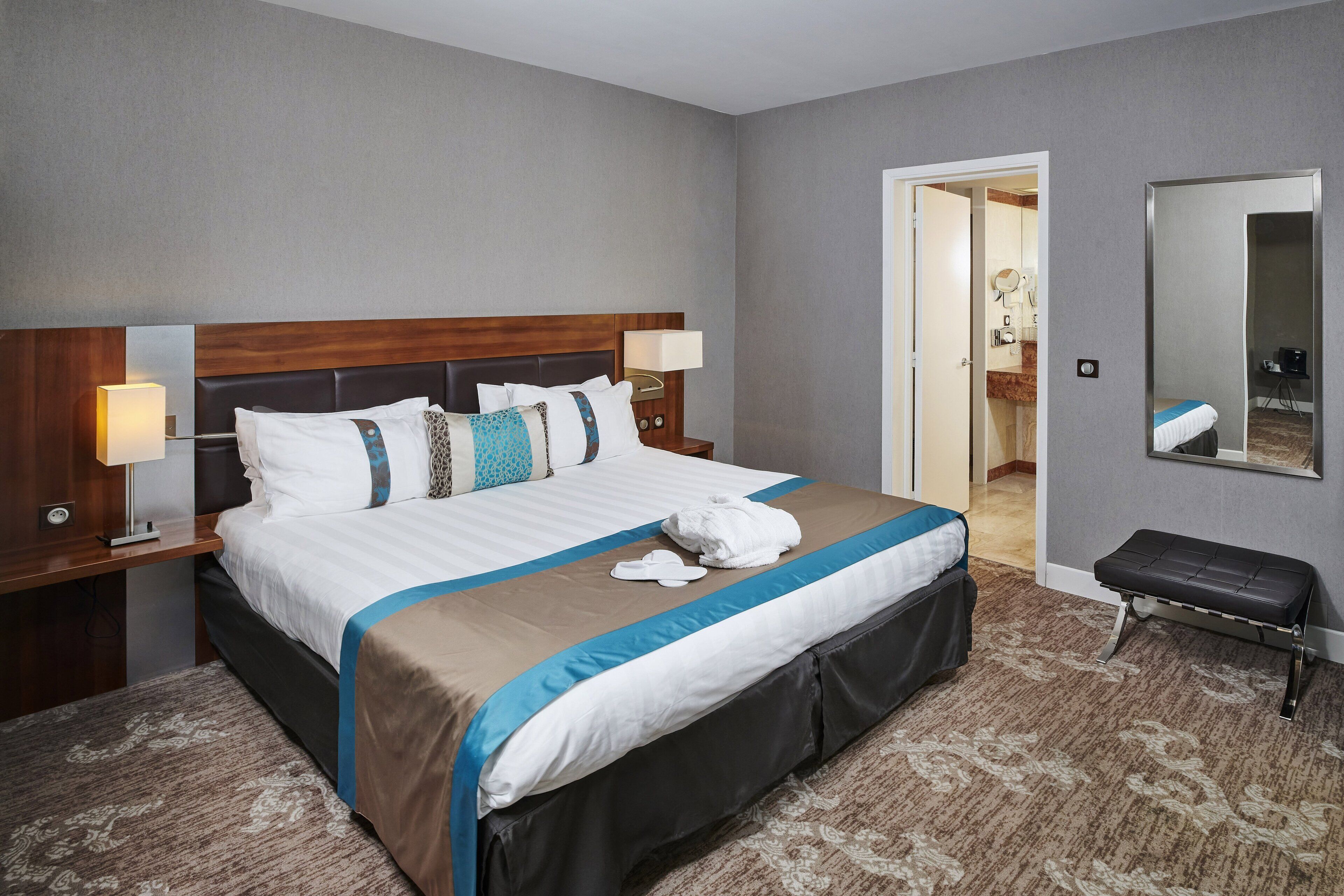 A hotel room with a large bed, a bedside table with a lamp, a bench, and a mirror on the wall.