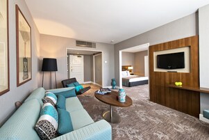 Habitación de hotel moderna con un sofá grande, una mesa redonda pequeña, un televisor de pantalla plana montado en la pared y una puerta que da al baño.