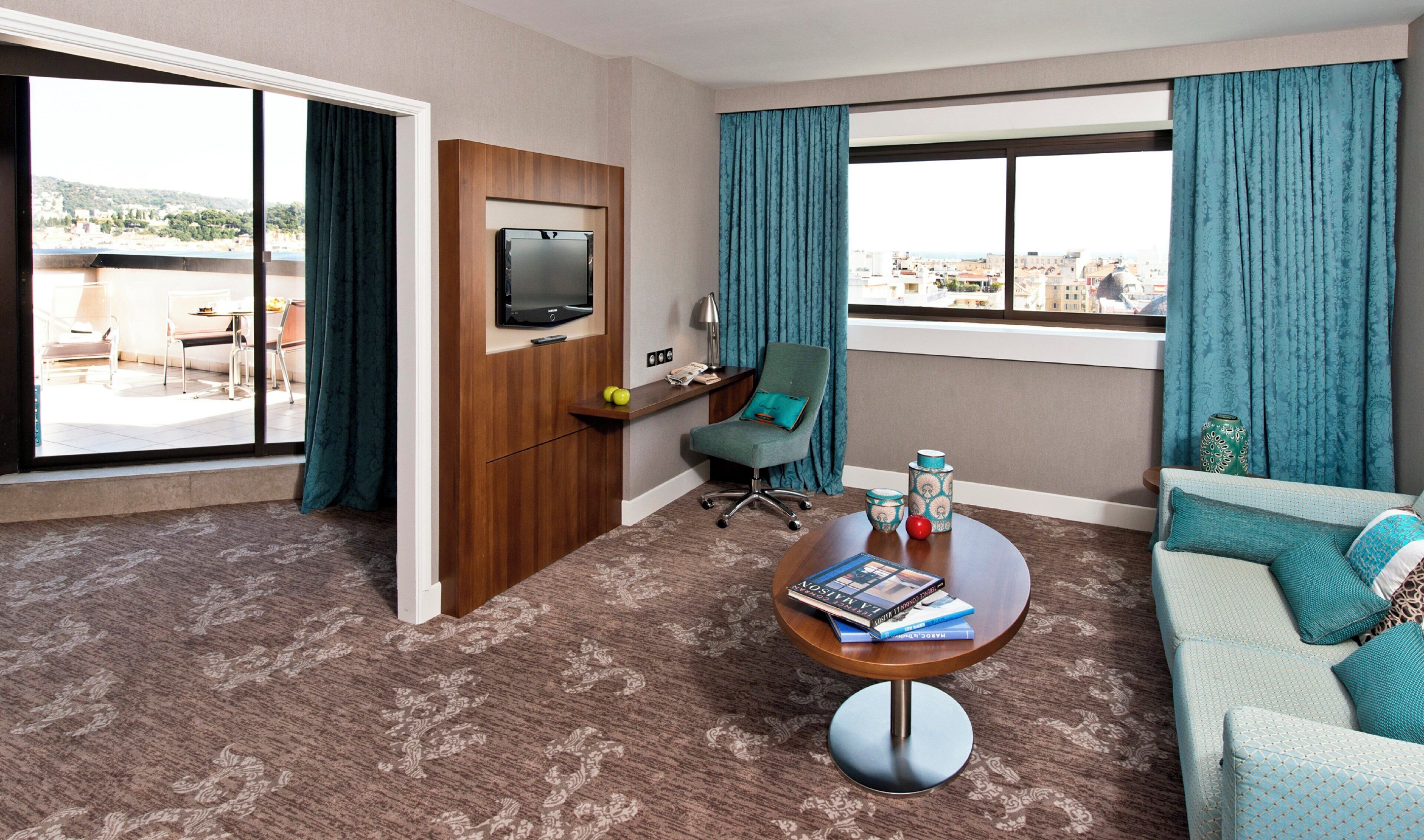Habitación de hotel con televisor, escritorio con lámpara, silla, sofá, mesa de centro con libros y vistas a la ciudad.