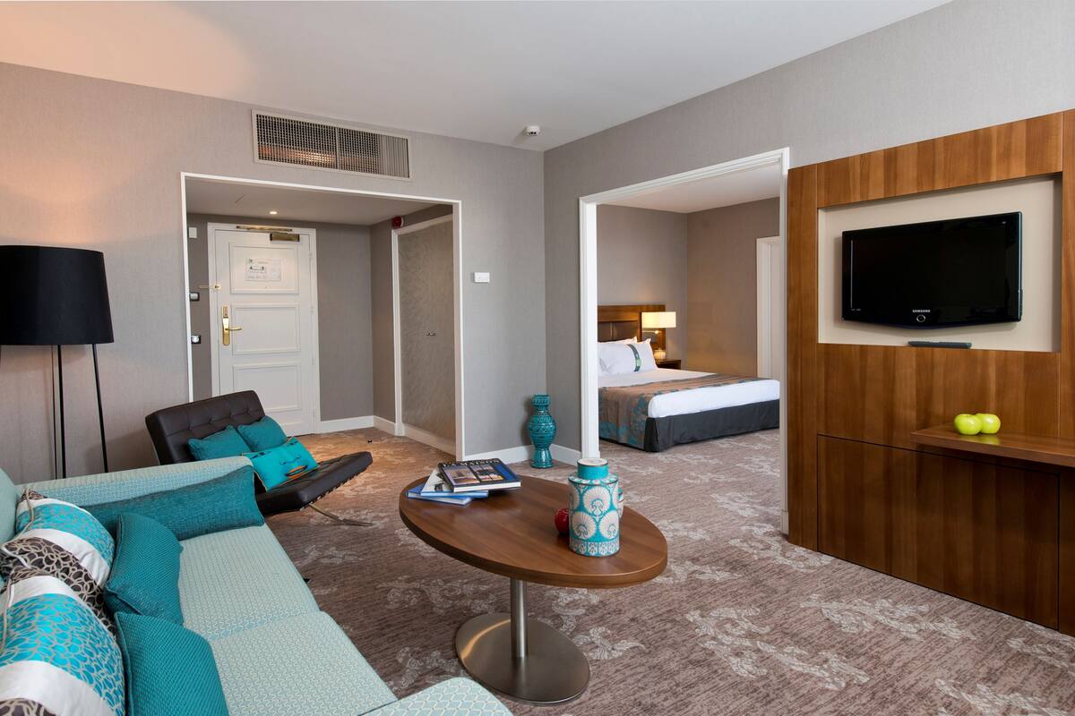 A hotel room with a sofa, a small table, a TV mounted on the wall, and a door leading to another room.