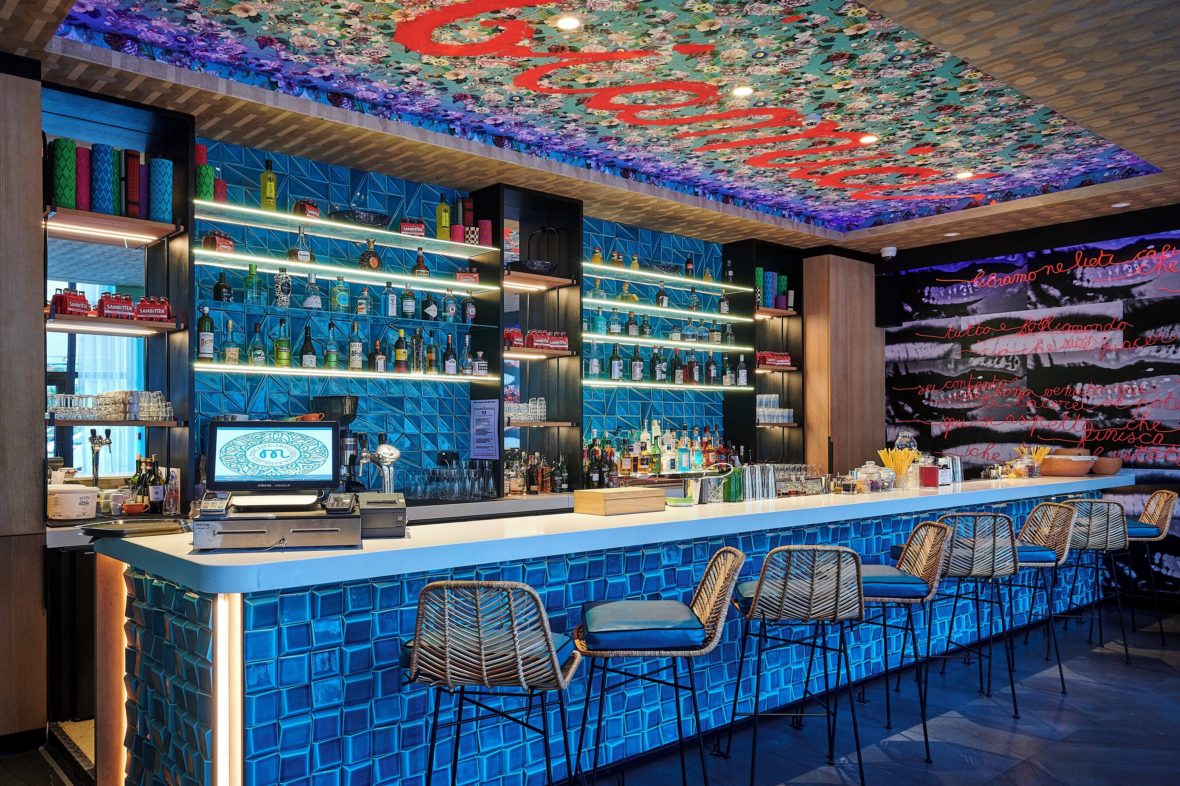 Un bar avec un mur recouvert de carreaux bleus, des étagères remplies de bouteilles d’alcool et des tabourets de bar.