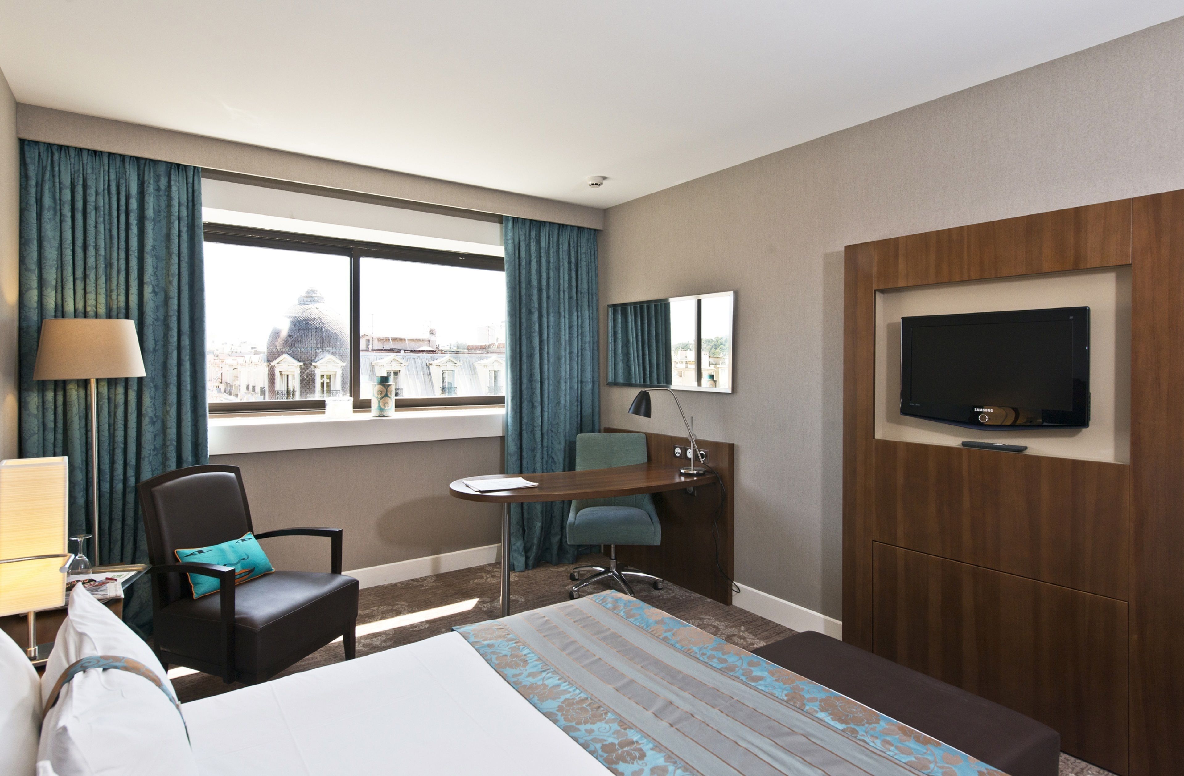 A hotel room with a bed, desk, chair, TV, and large window with curtains.