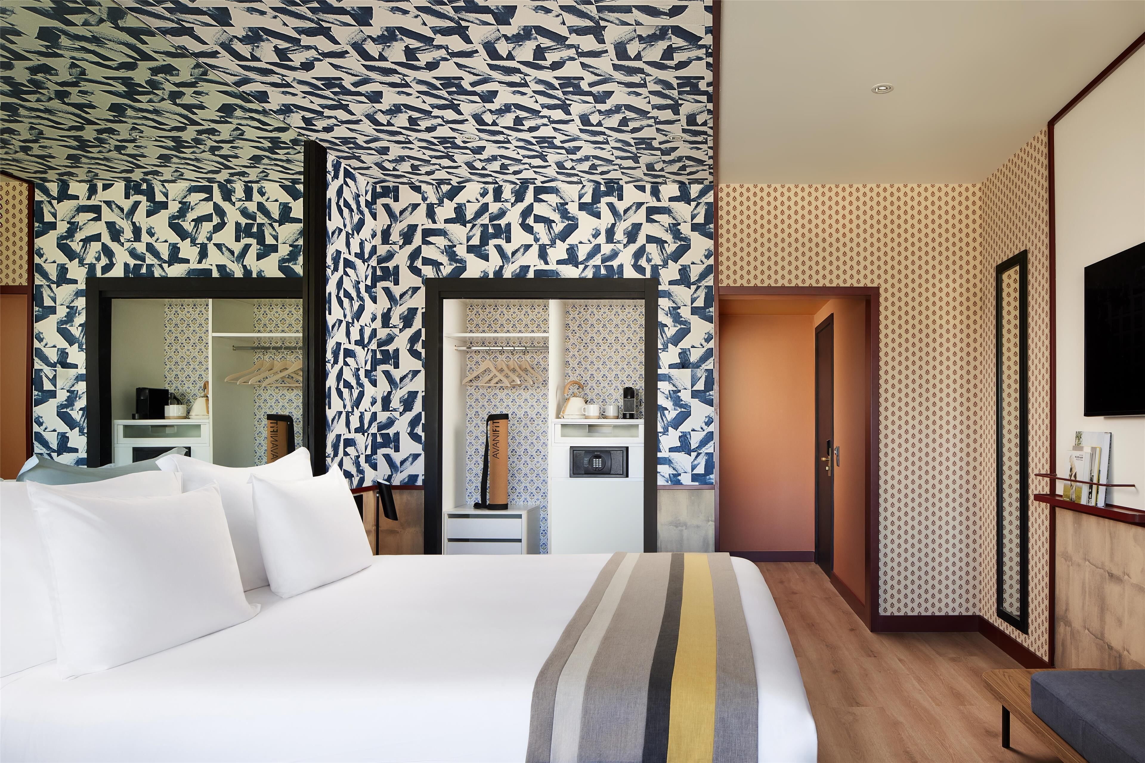 Une chambre d’hôtel comprenant un grand lit, une armoire, une coiffeuse avec un miroir, une petite salle de bain avec un lavabo et des toilettes, et un téléviseur fixé au mur.