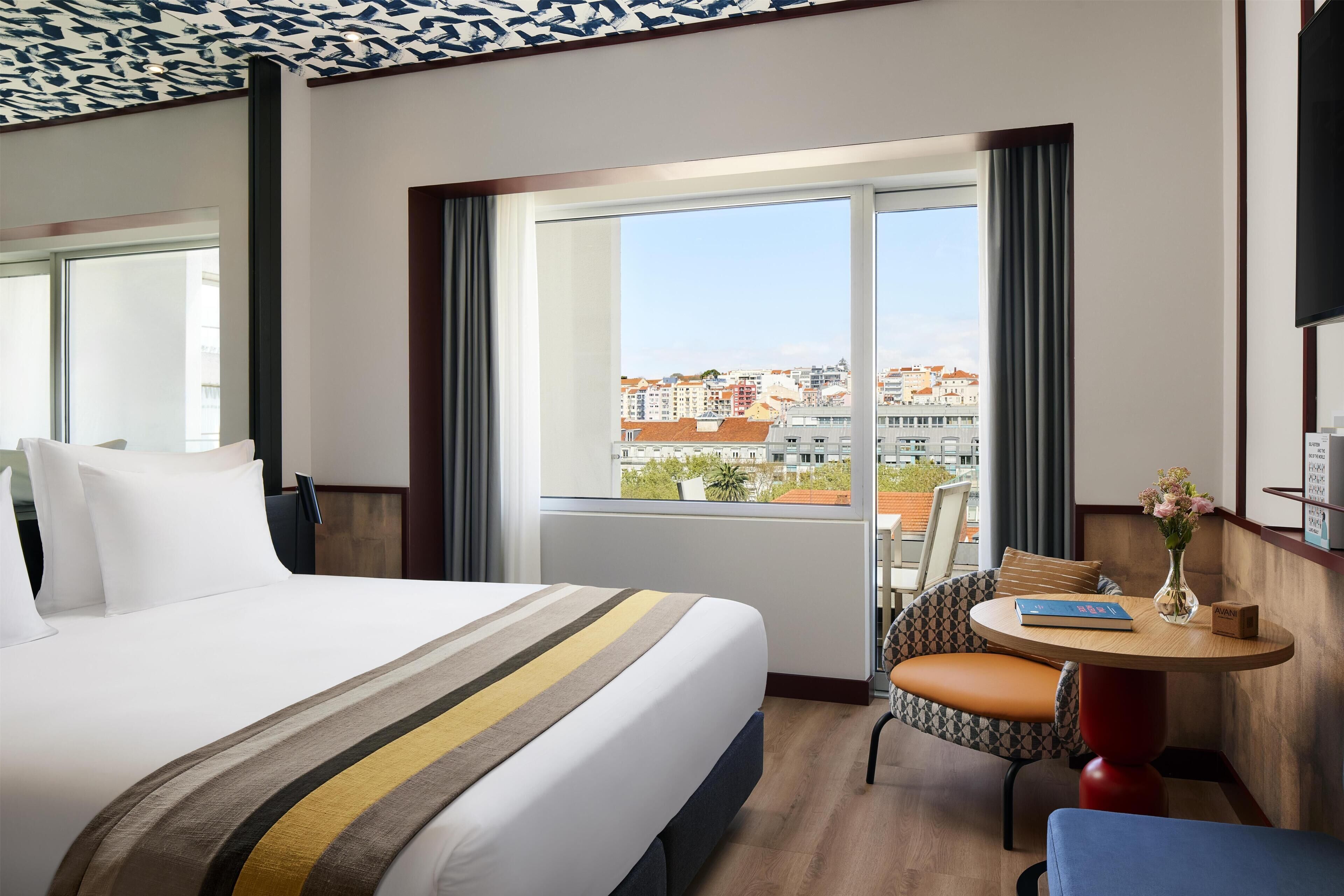 A hotel room with a large bed, a round table, a chair, and a view of the city through large windows.
