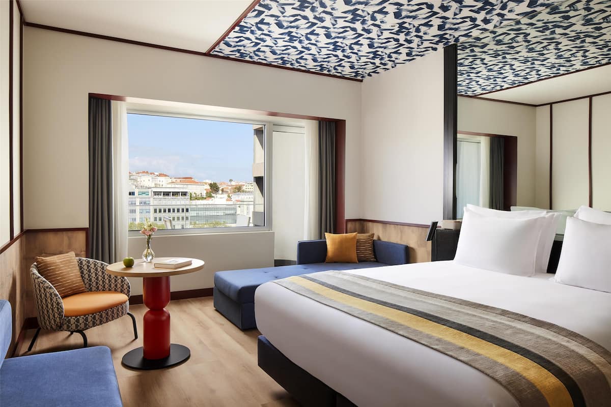 Une chambre d’hôtel avec un grand lit, un canapé, une petite table et une vue sur les bâtiments à travers la fenêtre.