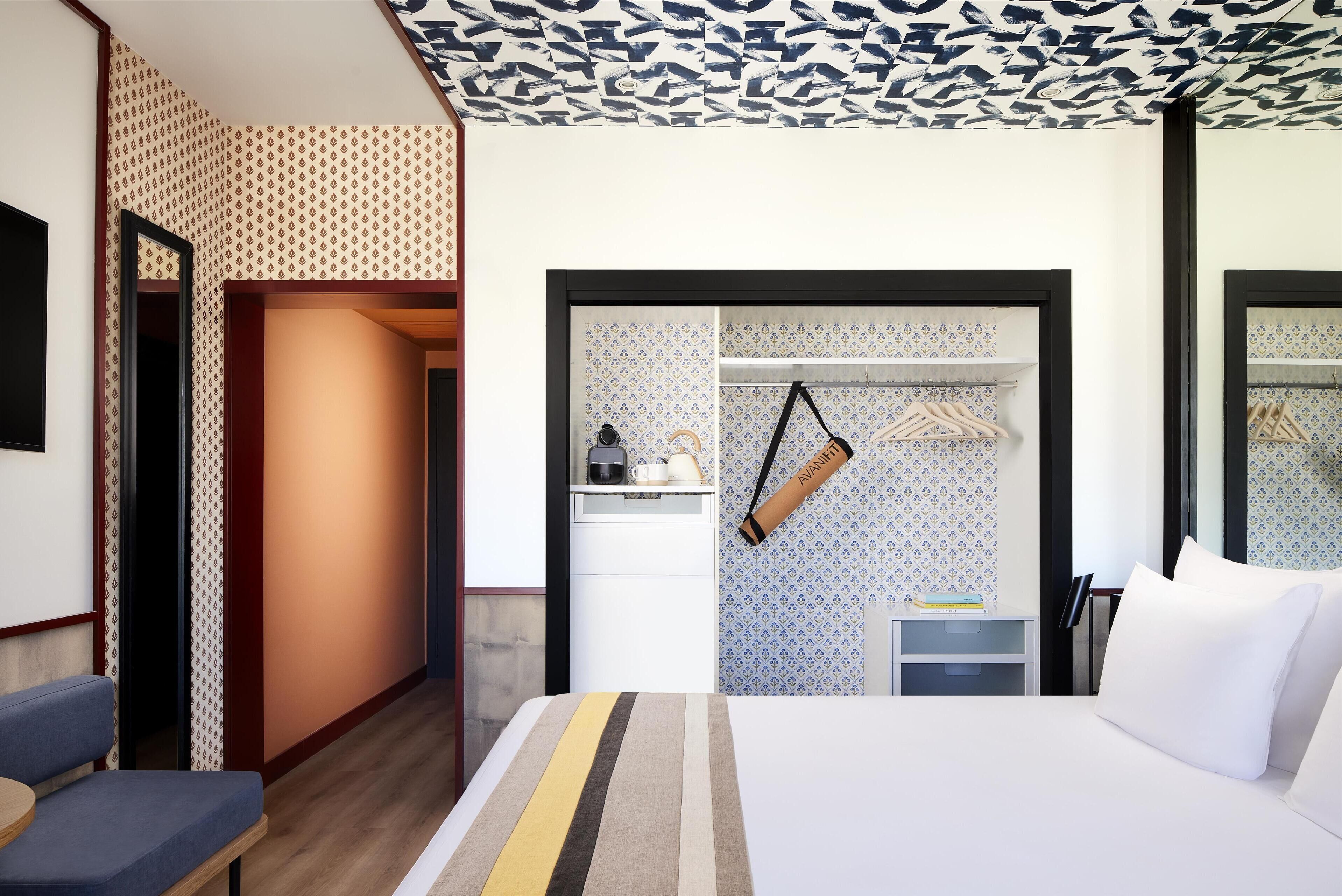 Quarto de hotel com cama, um banco, guarda-roupa e pequena área de cozinha.