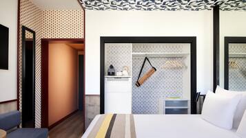 Quarto de hotel com cama, um banco, guarda-roupa e pequena área de cozinha.