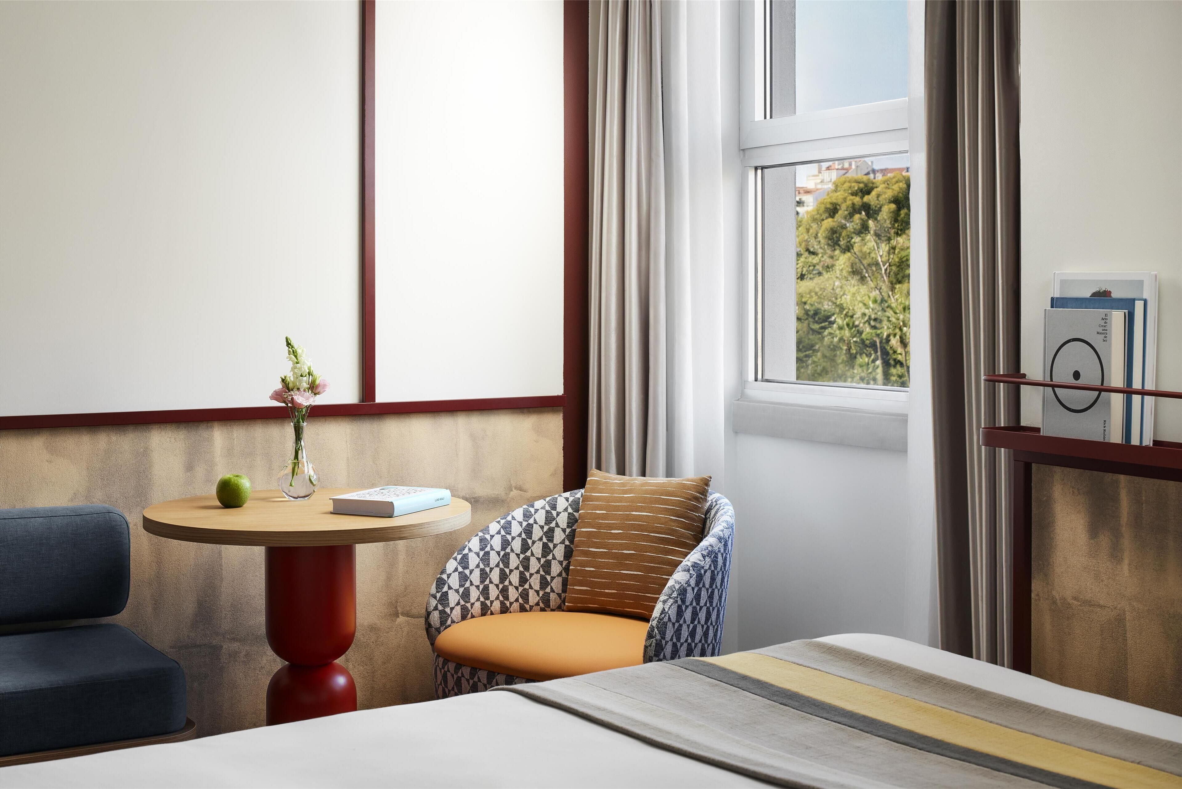 Ein modernes Zimmer mit einem runden Holztisch, einem gemusterten Sessel, einem blauen Sofa und einem Bett mit gestreifter Kopfteil.