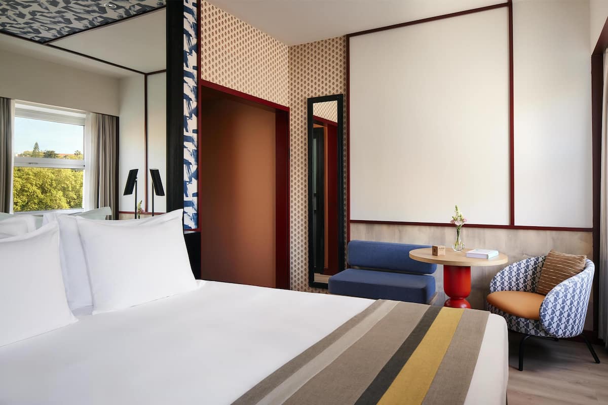 Une chambre d’hôtel avec un grand lit, une petite table ronde ornée d’un vase, un fauteuil bleu et un mur à motifs.