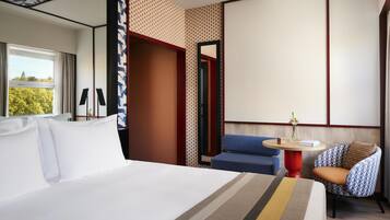 Camera d'albergo con un letto grande, un tavolino rotondo con un vaso, una poltrona blu e una parete decorata.