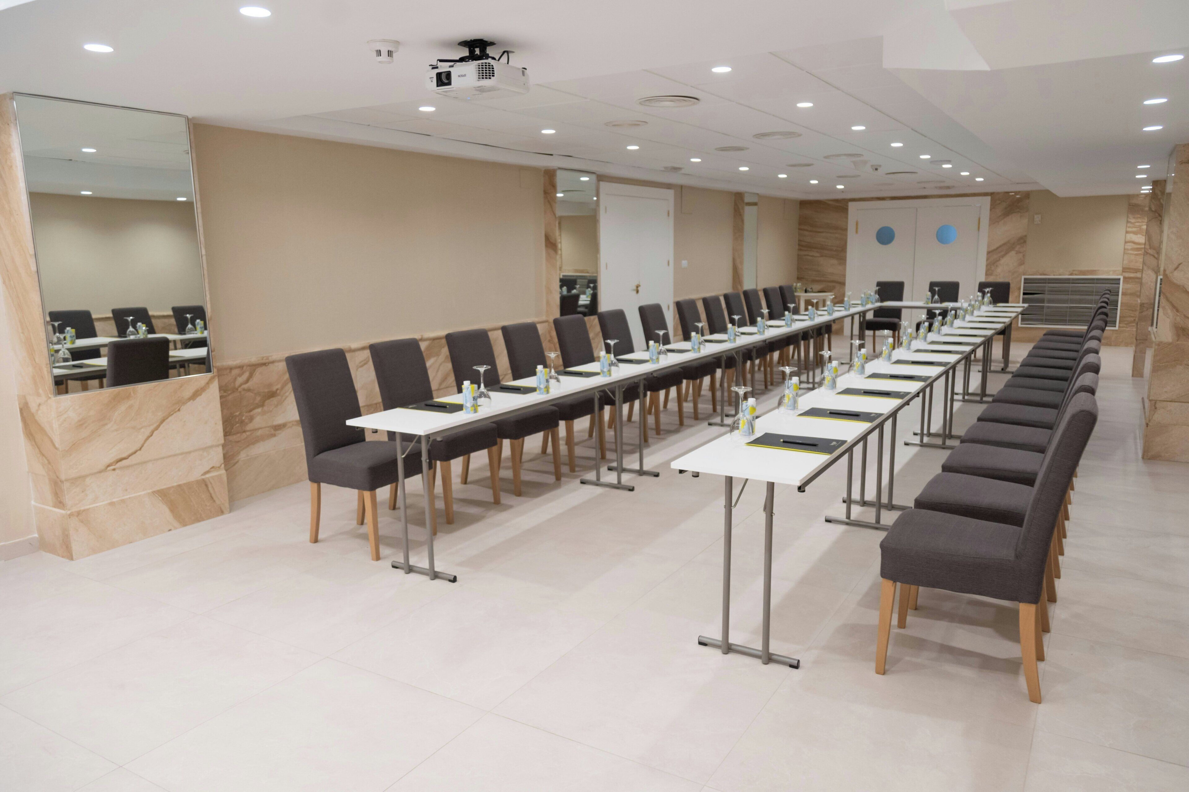 Een vergaderzaal met lange tafels, stoelen en een beamer.