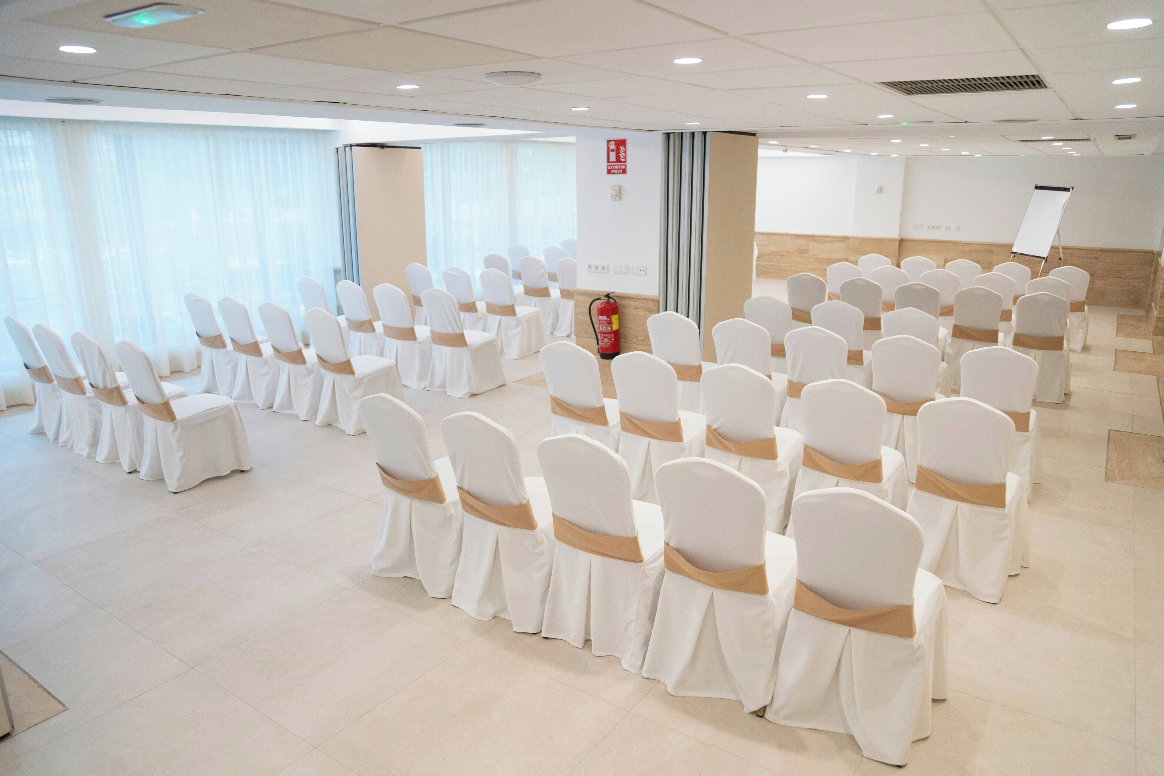 A conference room with rows of white chairs with brown sashes, a whiteboard, and a fire extinguisher.