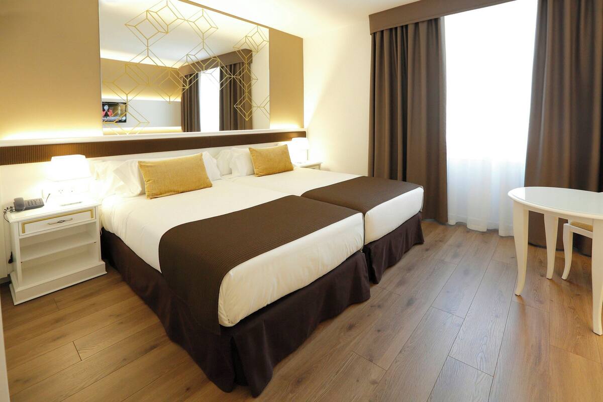 Habitación de hotel con una cama grande, mesitas de noche, un escritorio, un espejo y vistas al exterior.