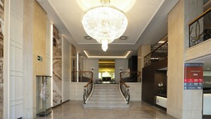 A hotel lobby with a grand chandelier, marble columns, a reception desk, and a staircase with metal railings.