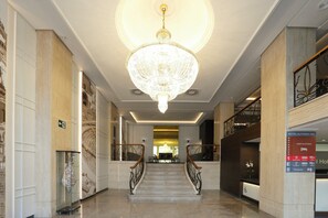Eine Hotelhalle mit einem prunkvollen Kronleuchter, Marmorsäulen, einem Empfangstresen und einer Treppe mit Metallgeländern.