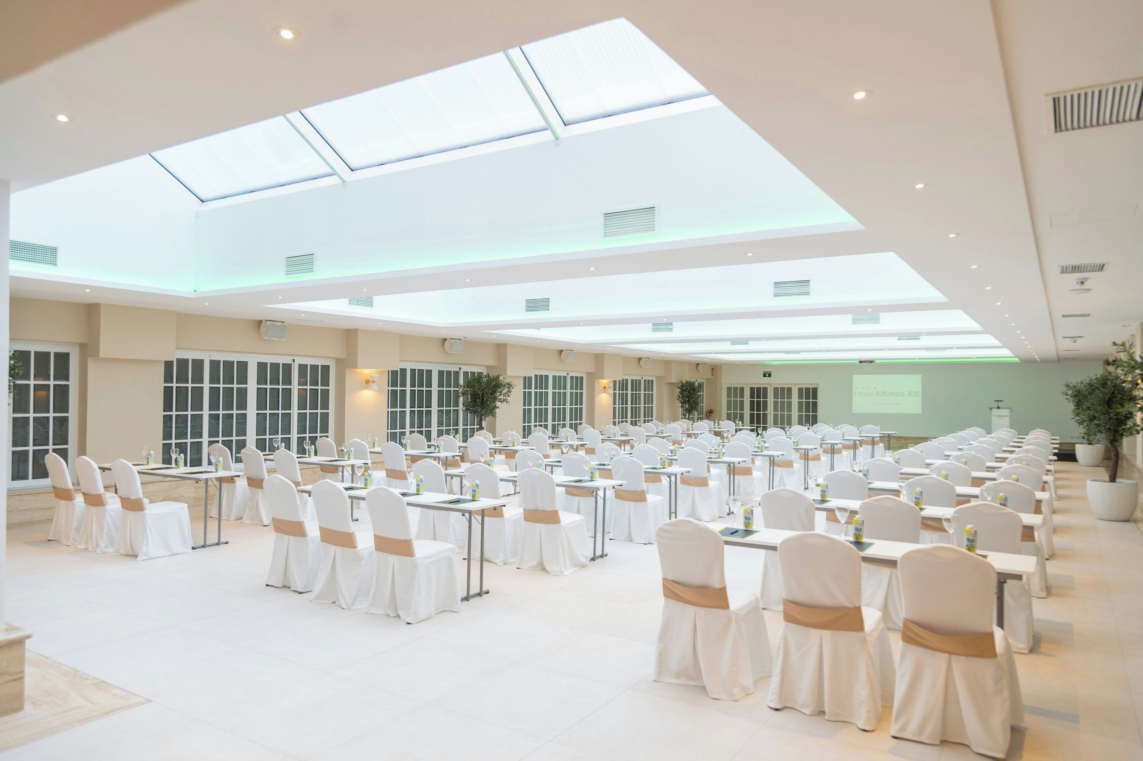 A spacious indoor venue with rows of tables and chairs, large windows, and a presentation screen.