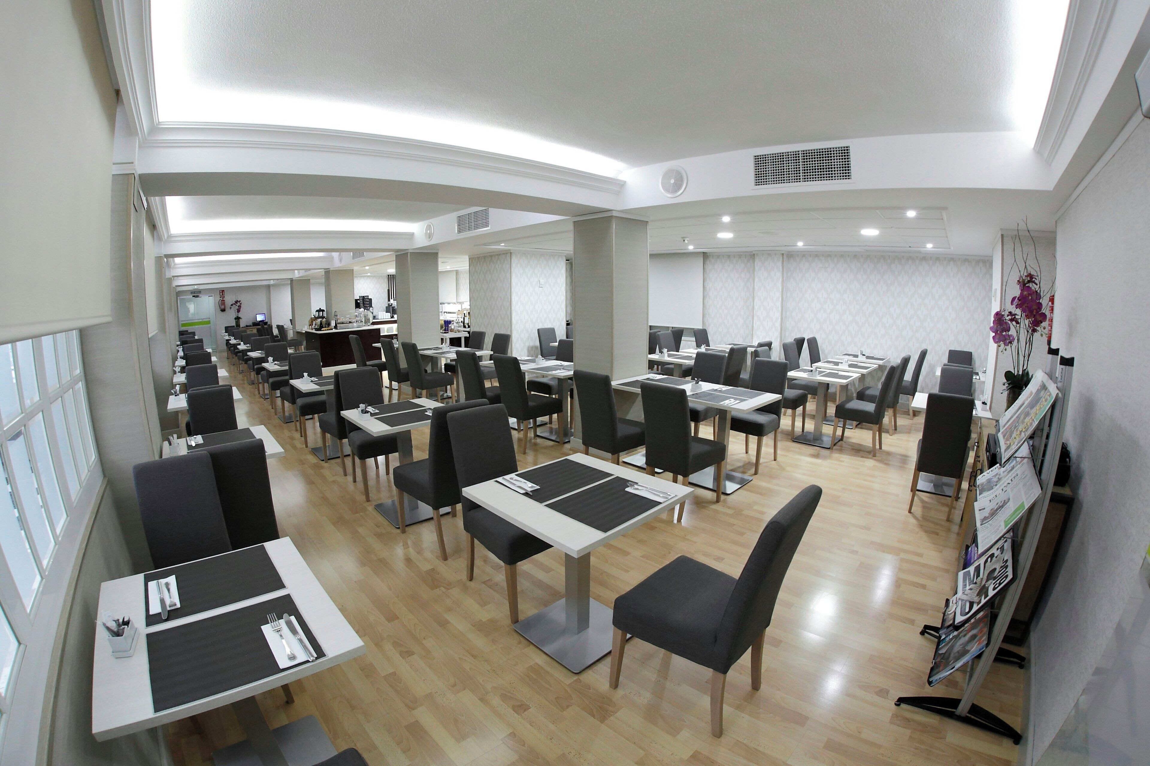 Amplia zona de comedor de hotel con varias mesas y sillas, un mostrador de recepción y vistas al vestíbulo.