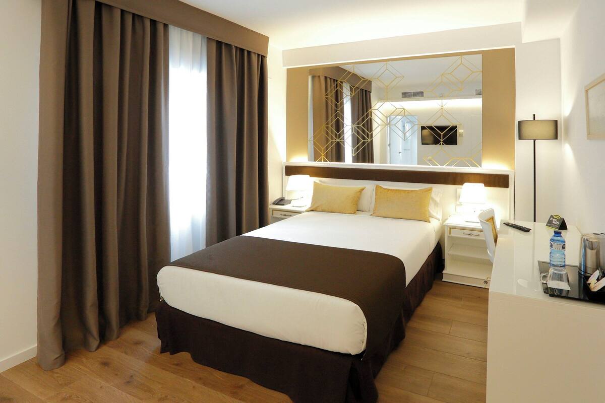 Habitación de hotel con una cama grande, una mesita de noche con lámpara, un espejo con diseños geométricos y otra mesita.