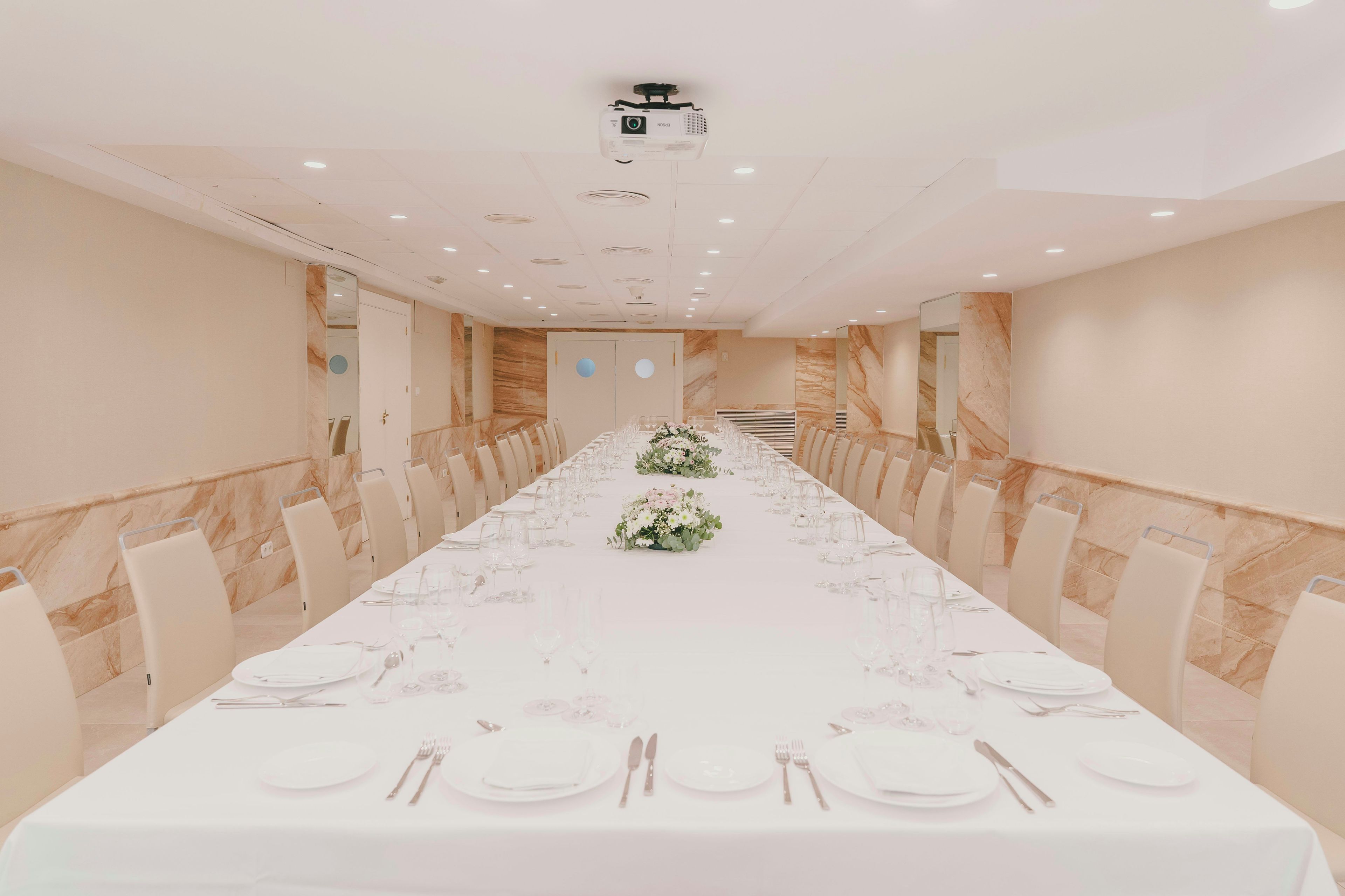 Un salón de conferencias con una larga mesa dispuesta para un evento formal, equipado con un proyector y un espejo grande en la pared.
