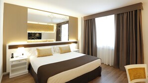 A hotel room with a large bed, a bedside table with a lamp, a chair, a large mirror, and a window with curtains.