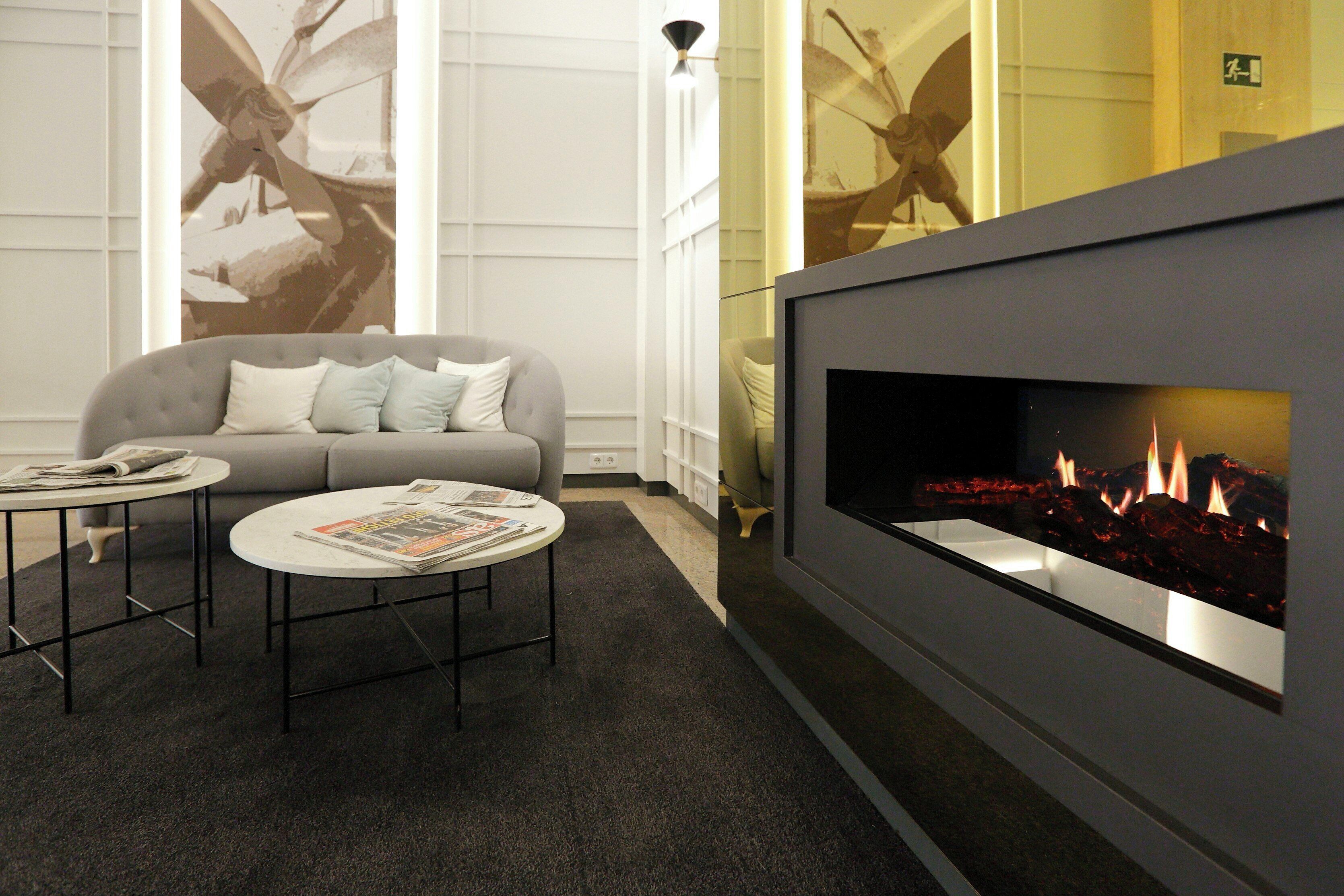 A modern lobby with a grey sofa, two round tables, a fireplace, and large windows with artwork.