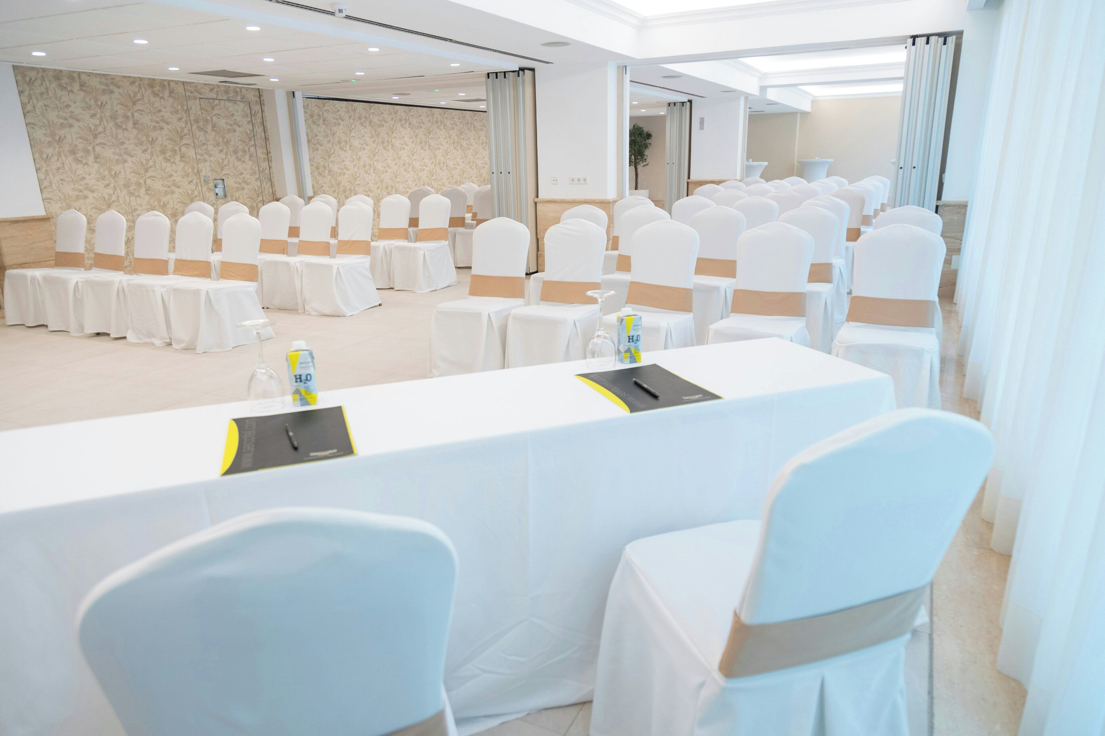 Une salle de conférence avec des rangées de chaises et de tables blanches, des murs de couleur beige et une palette de couleurs neutres.