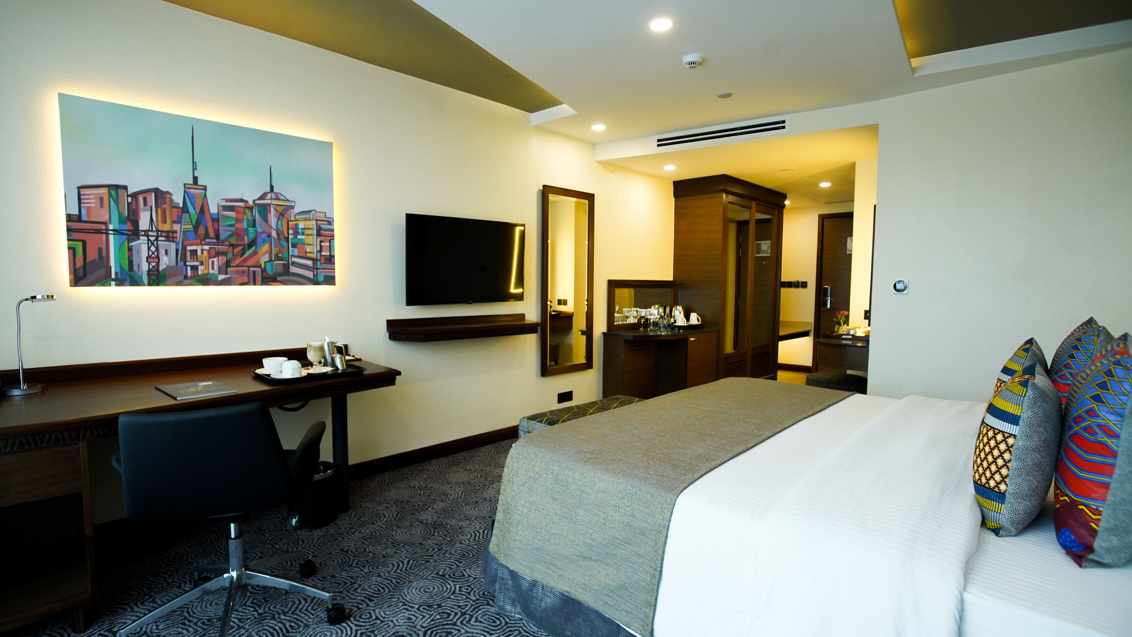 A hotel room with a bed, a desk, a chair, a TV, a mirror, and a painting on the wall.