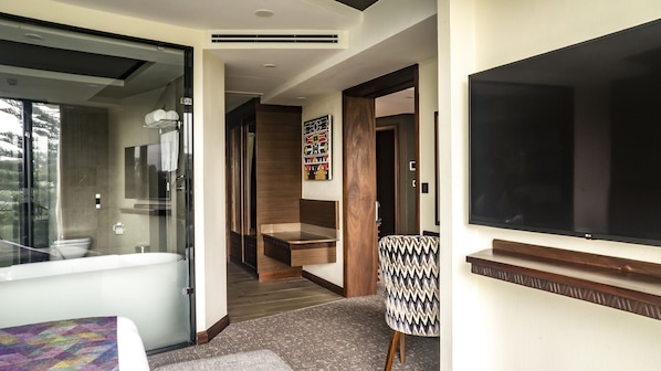A modern hotel room with a glass shower enclosure, a wooden staircase, and a large flat-screen TV.