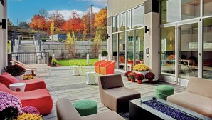 An outdoor patio with modern seating, a fire feature, and a view of autumn trees. - Aloft by Marriott Framingham (Framingham)
