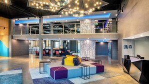 A spacious lobby with a modern design, featuring a central seating area with colorful cushions, a reception desk, and a mezzanine level with seating and a bar area. - Aloft by Marriott Framingham (Framingham)