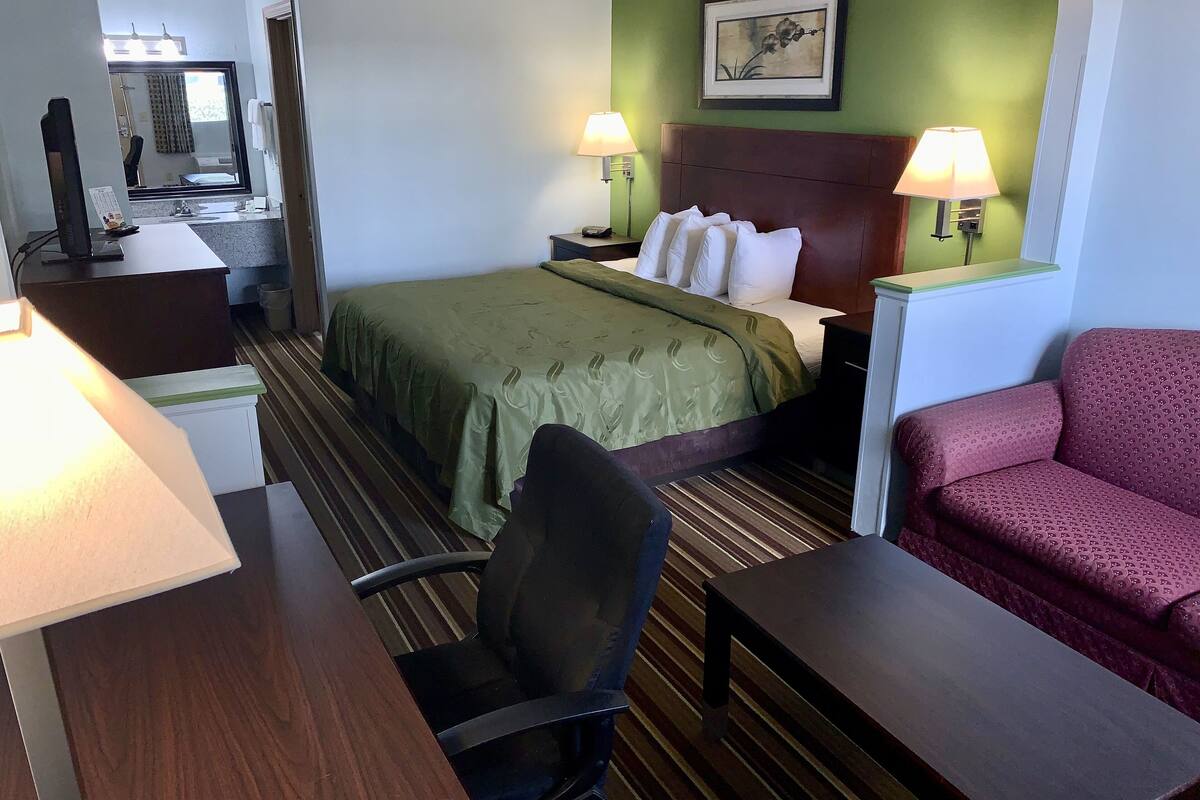 A hotel room with a bed, two bedside lamps, a desk with a chair, a red armchair, and a large mirror.