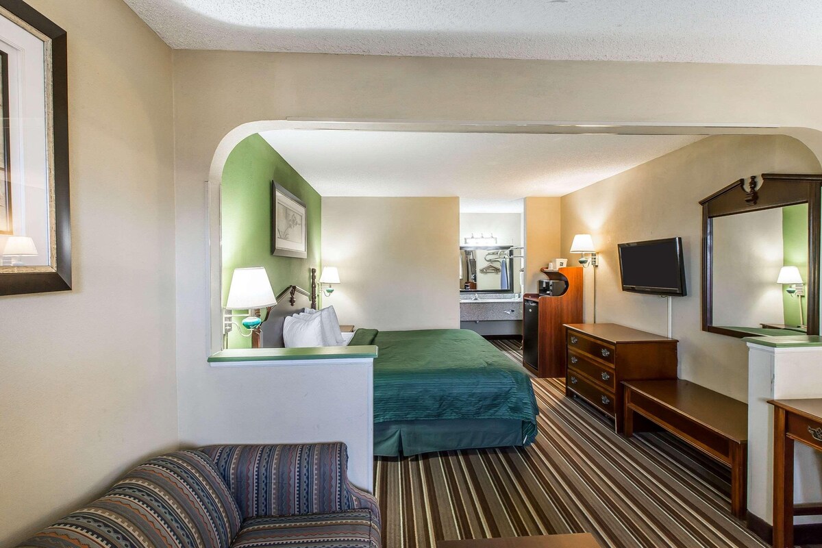 A hotel room with a bed, a desk, a chair, a television, and a bathroom visible in the background.