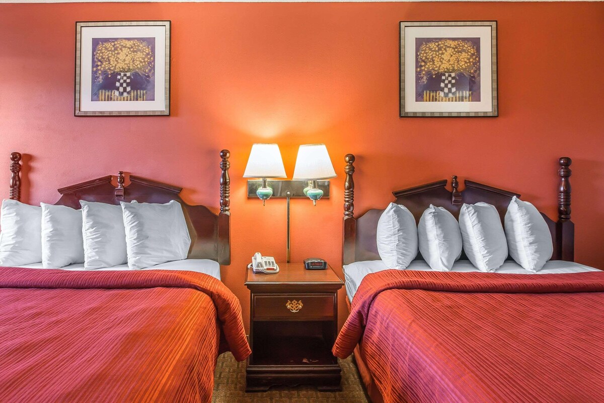 A hotel room with two beds, each with white linens and red bedding. There are two wall-mounted lamps with green shades and a small table between the beds.