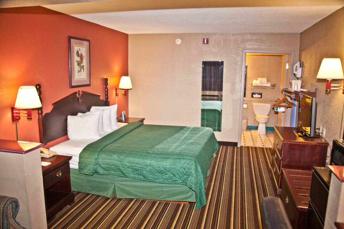 A hotel room with a bed, bedside tables, lamps, a mirror, a dresser, and a bathroom visible through an open door.