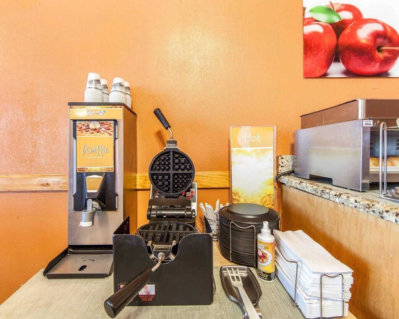A waffle maker, a waffle dispenser, a waffle iron, a stack of napkins, a bottle of syrup, and a microwave in a kitchen area.