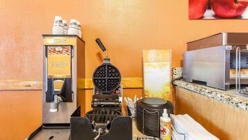 A waffle maker, a waffle dispenser, a waffle iron, a stack of napkins, a bottle of syrup, and a microwave in a kitchen area.