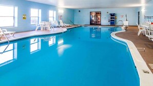 An indoor swimming pool with clear blue water, surrounded by white lounge chairs and a sunlit room with a door leading to another area.