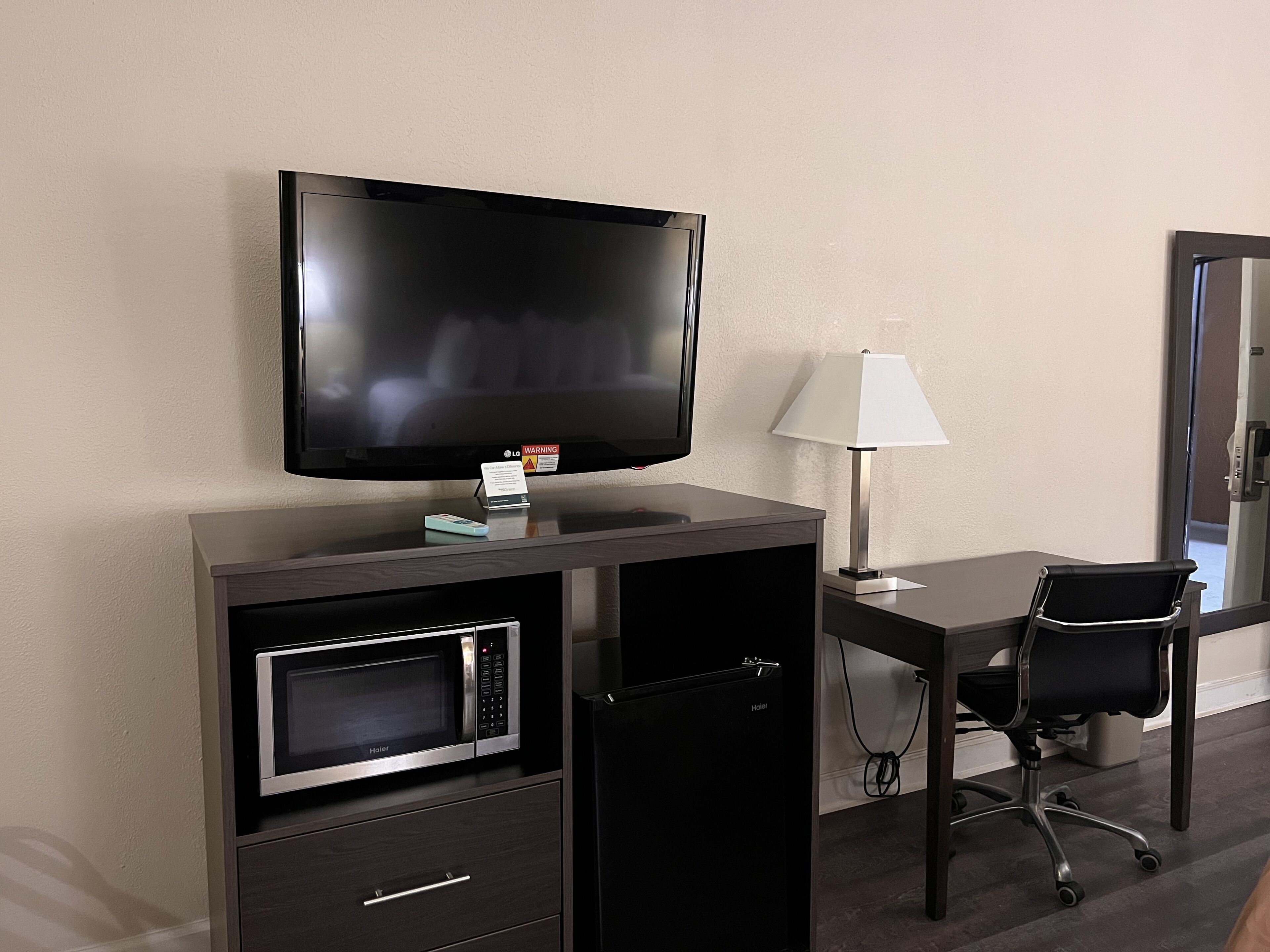 Quarto de hotel compacto com televisão de tela plana, micro-ondas, escrivaninha com luminária e uma cadeira.