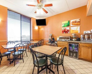 A dining area with tables and chairs, a ceiling fan, a TV mounted on the wall, and a kitchen counter with various food items.