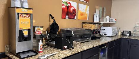 A breakfast bar with waffle and syrup dispensers, a waffle maker, a toaster, and a coffee machine.