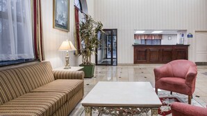 Un hall de hotel con un sofá, una mesa, una planta en maceta, un mostrador de recepción y una puerta que da a otra habitación.
