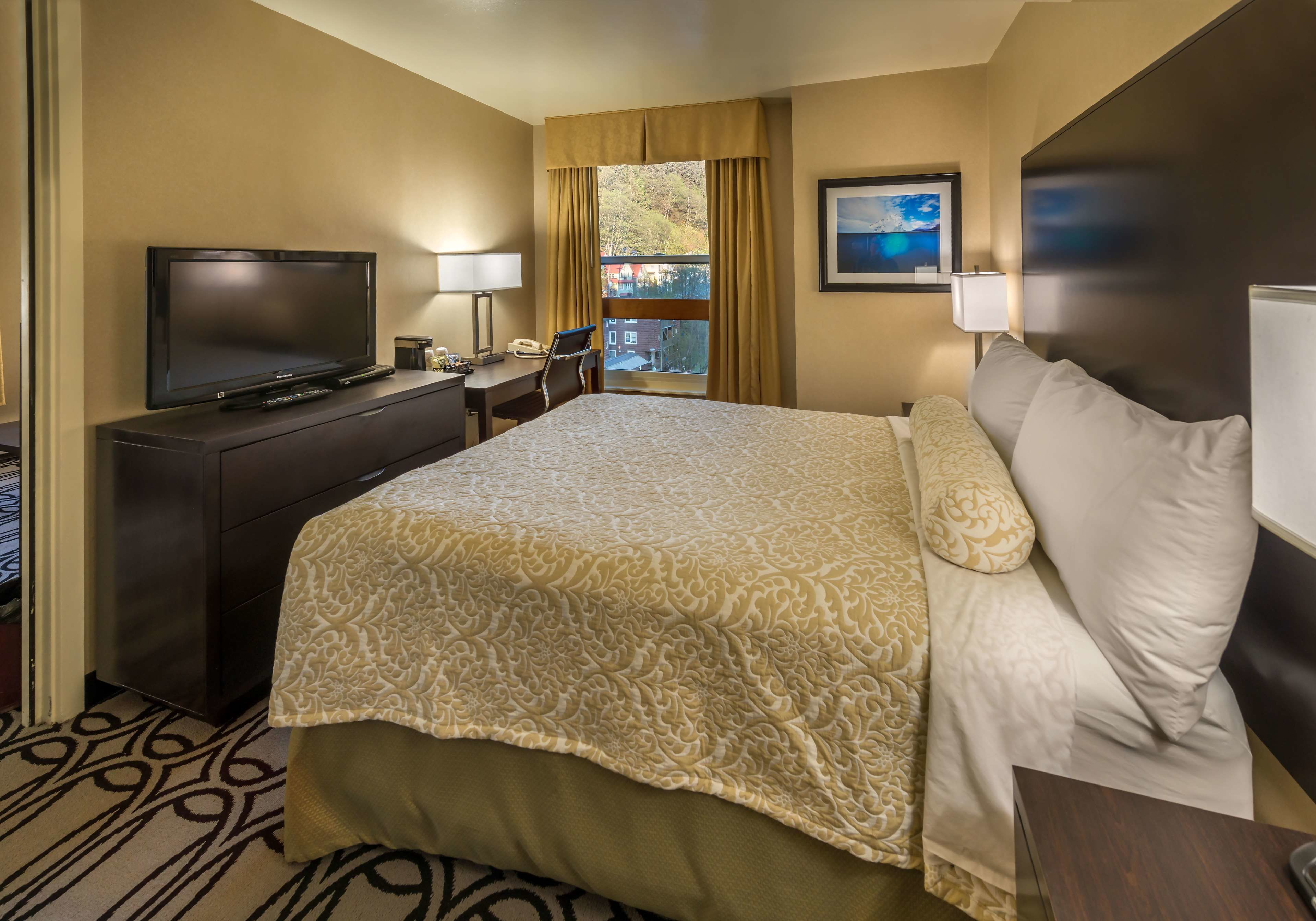 A hotel room with a large bed, a TV, a desk with a chair, and a window with curtains.
