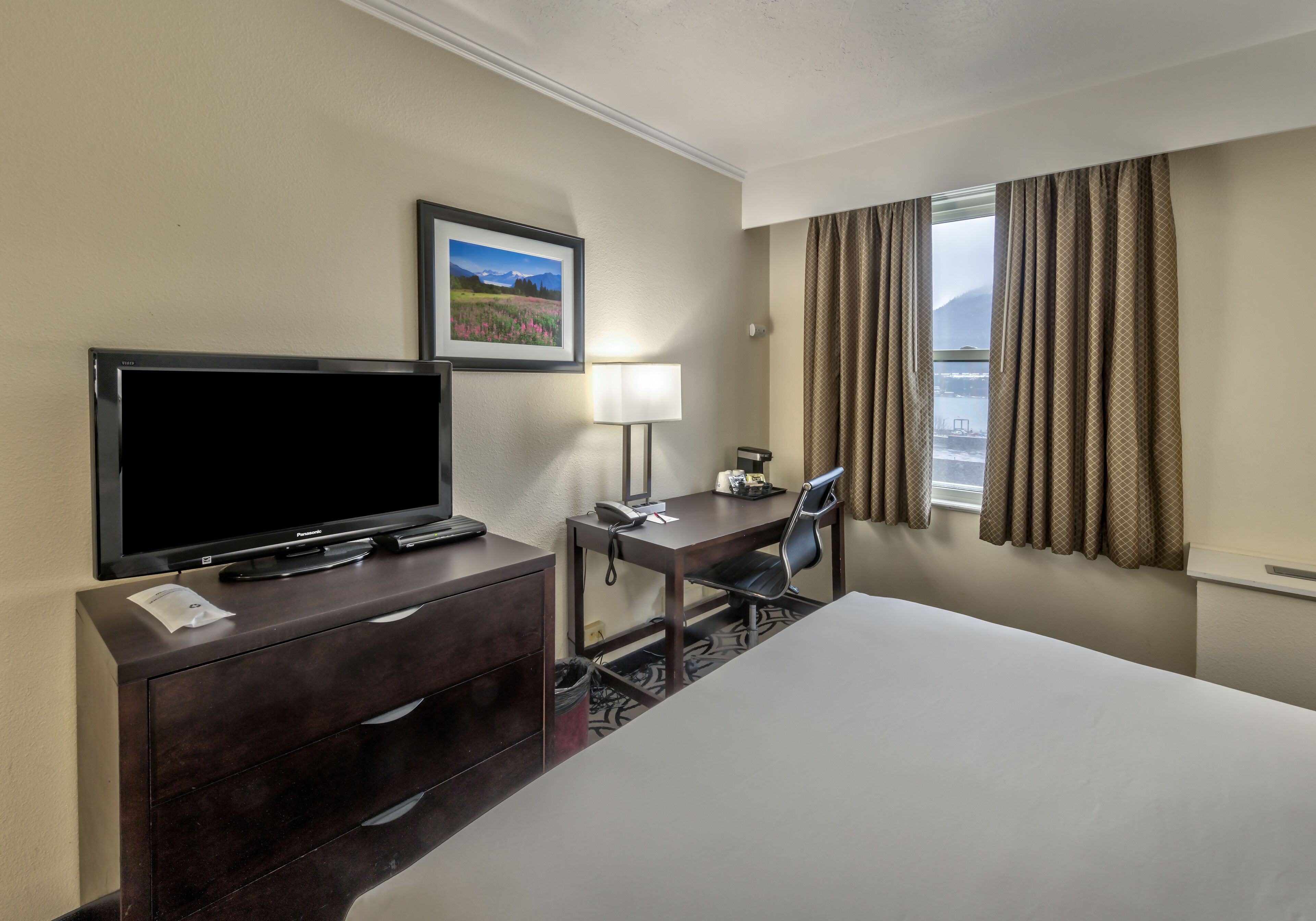 Hotelkamer met een bed, een bureau met stoel, een televisie, een lamp, een ingelijst schilderij aan de muur en een raam met gordijnen.