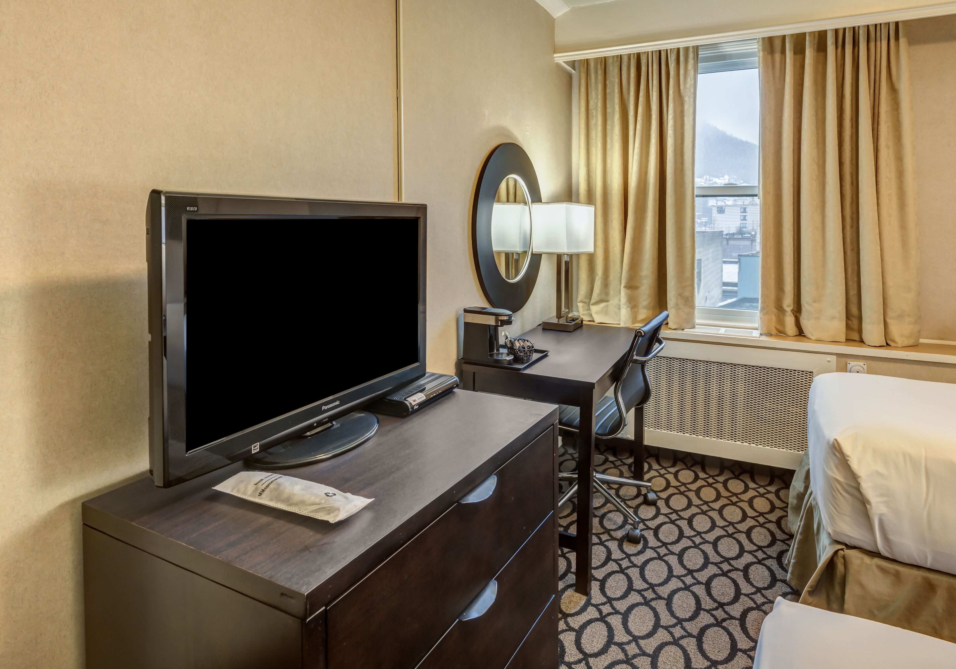 Une chambre d’hôtel équipée d’un téléviseur à écran plat, d’un bureau avec une lampe, d’une chaise, d’un lit et d’une fenêtre avec des rideaux.