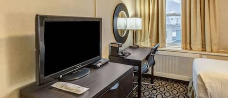 Quarto de hotel com televisão de tela plana, uma escrivaninha com luminária, uma cadeira, uma cama e uma janela com cortinas.