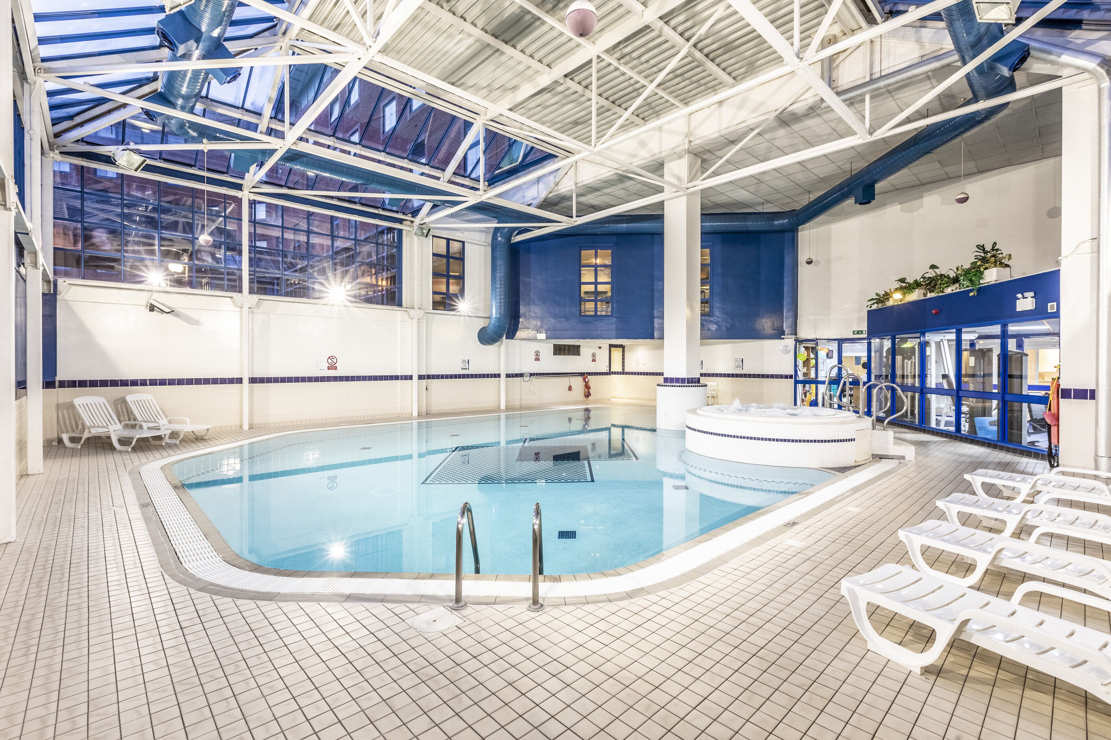An indoor swimming pool with clear blue water, surrounded by white lounge chairs and a high ceiling with skylights.