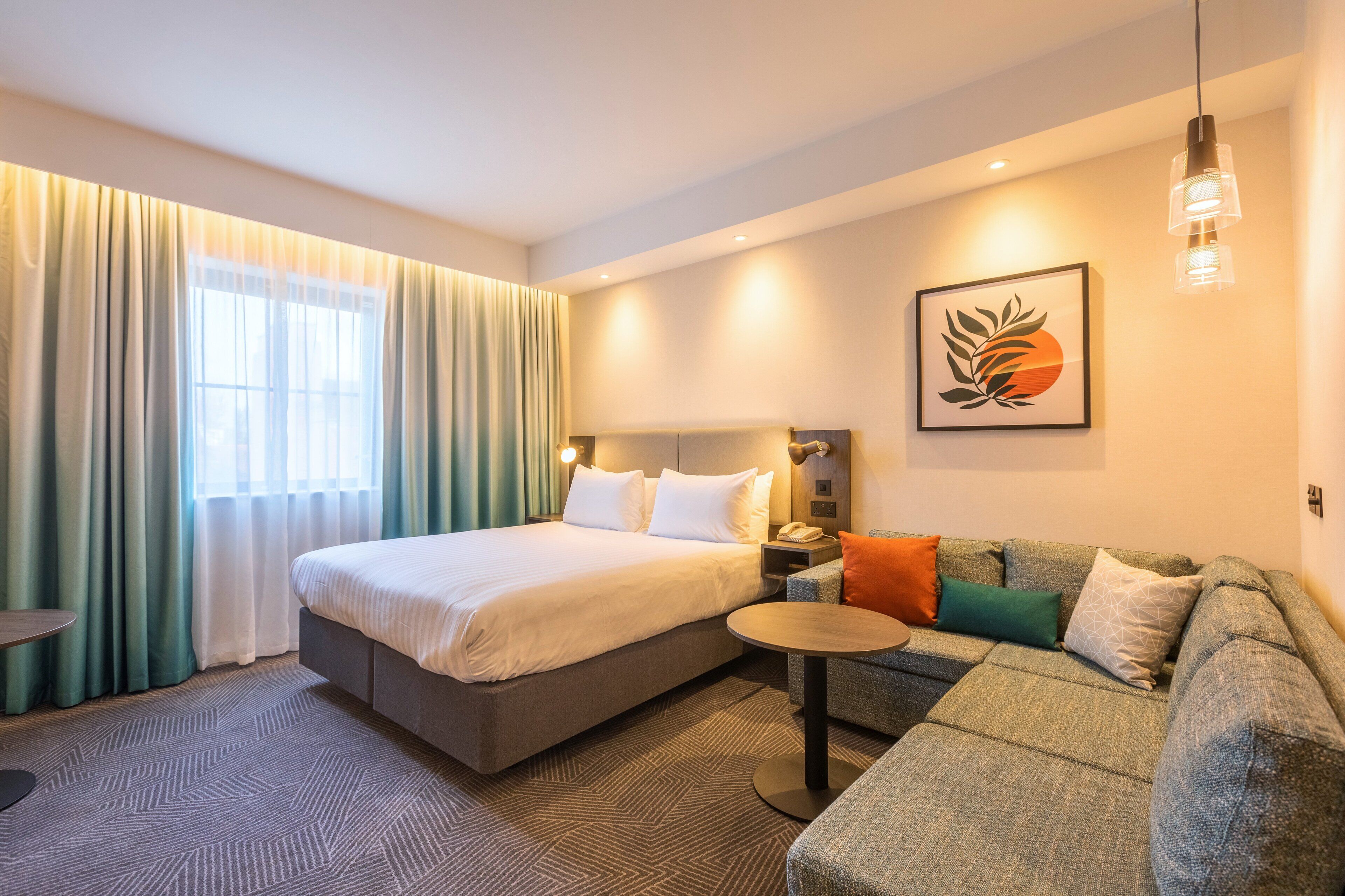 A hotel room with a bed, a sofa, a small table, and a painting on the wall.