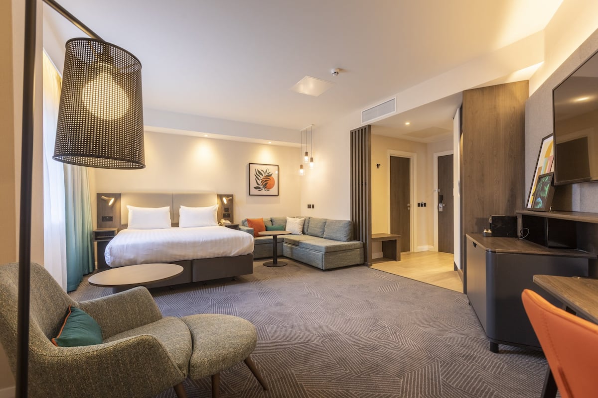 A modern hotel room with a bed, sofa, armchair, and a small table. There is a TV and a bookshelf.