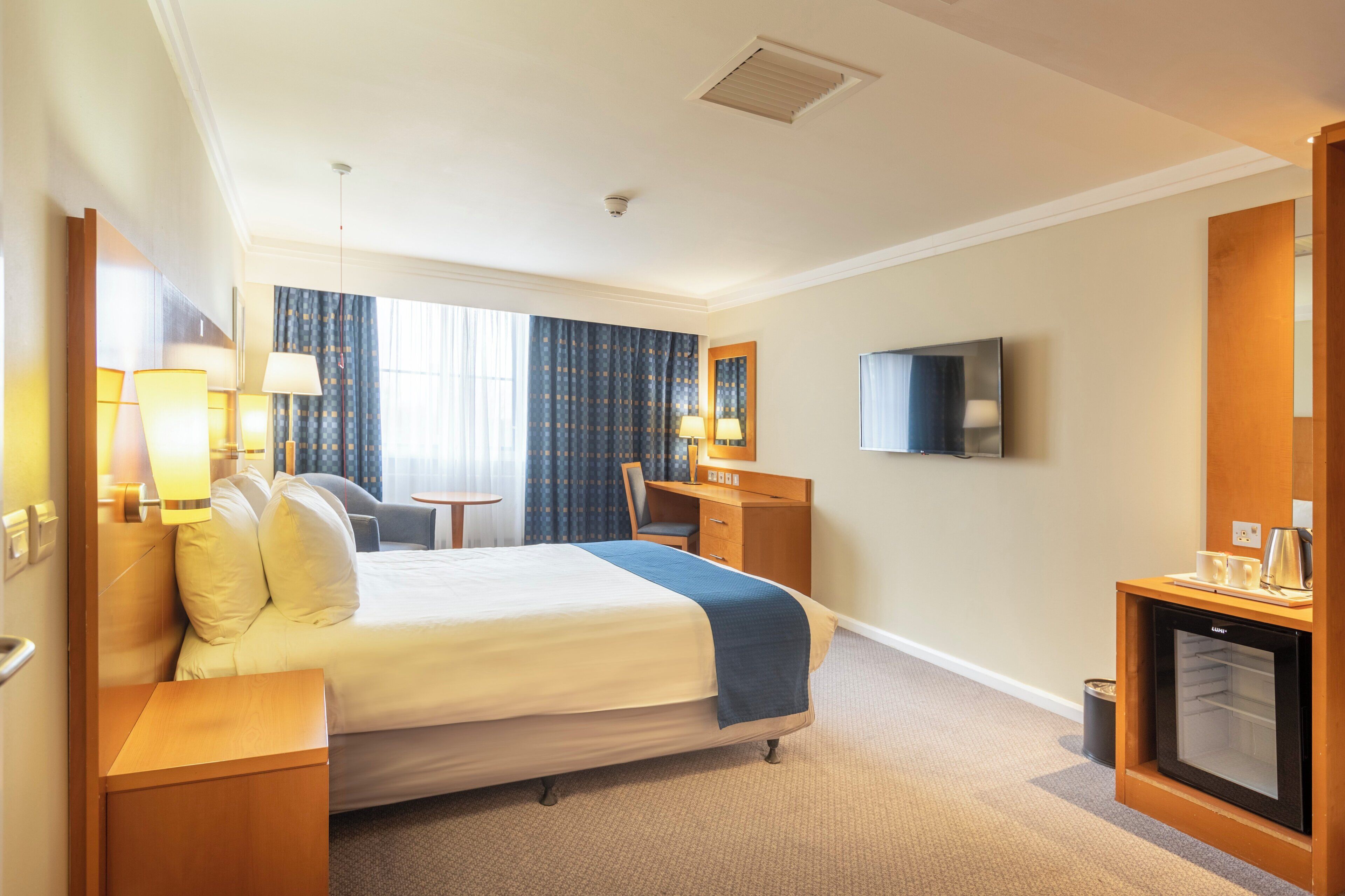 Una habitación de hotel con una cama, mesitas de noche, un escritorio, una silla, un televisor y un minibar.
